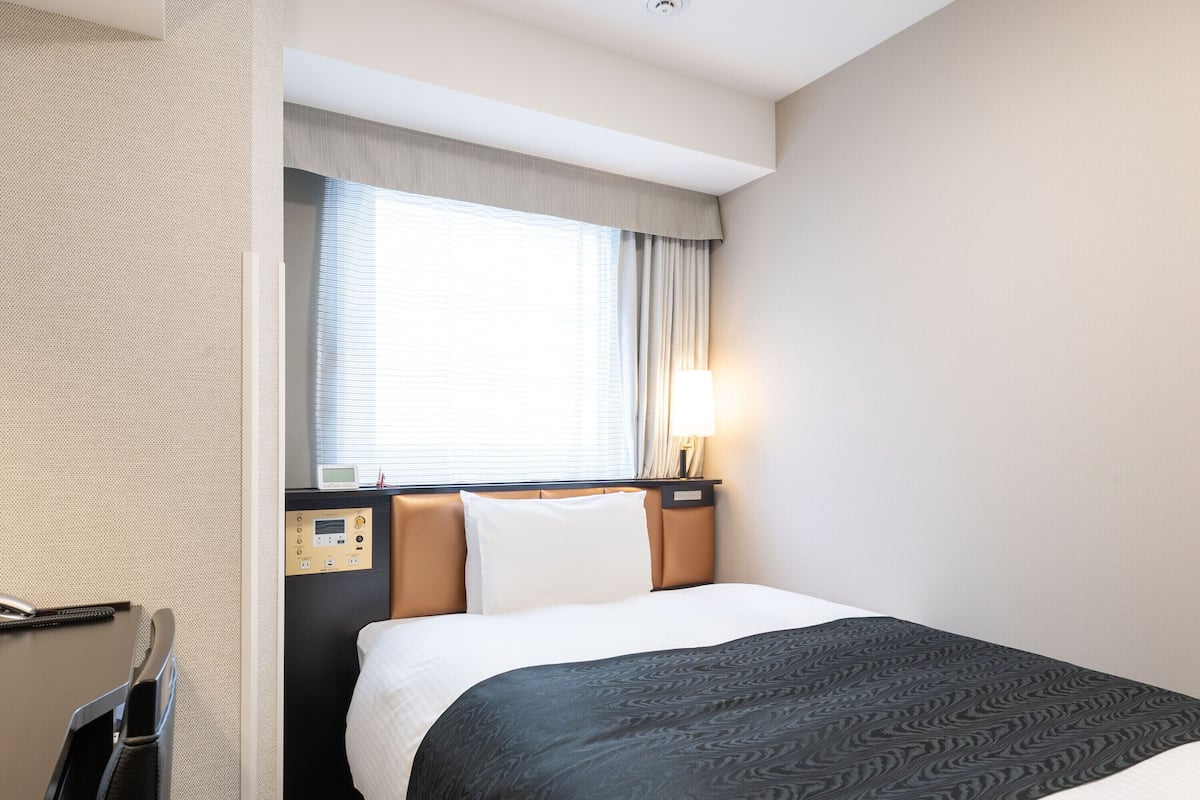 A hotel room with a bed, a bedside table, a lamp, a window with curtains, and a wall-mounted control panel.