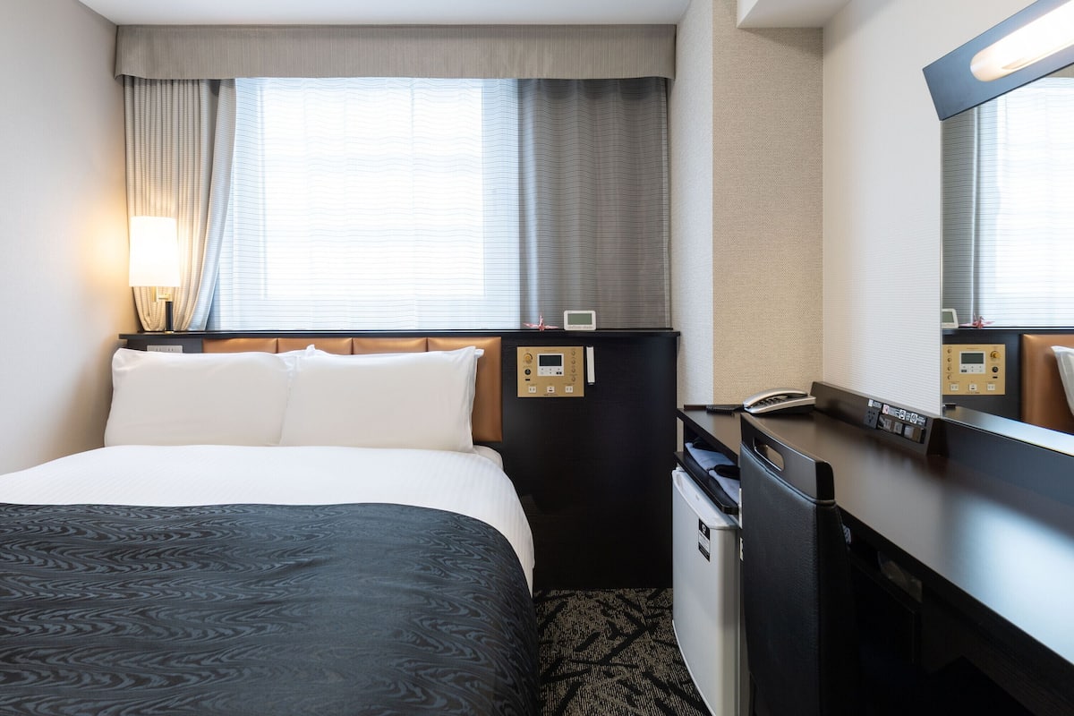 A hotel room with a bed, a desk, a mirror, a lamp, and a window with curtains.