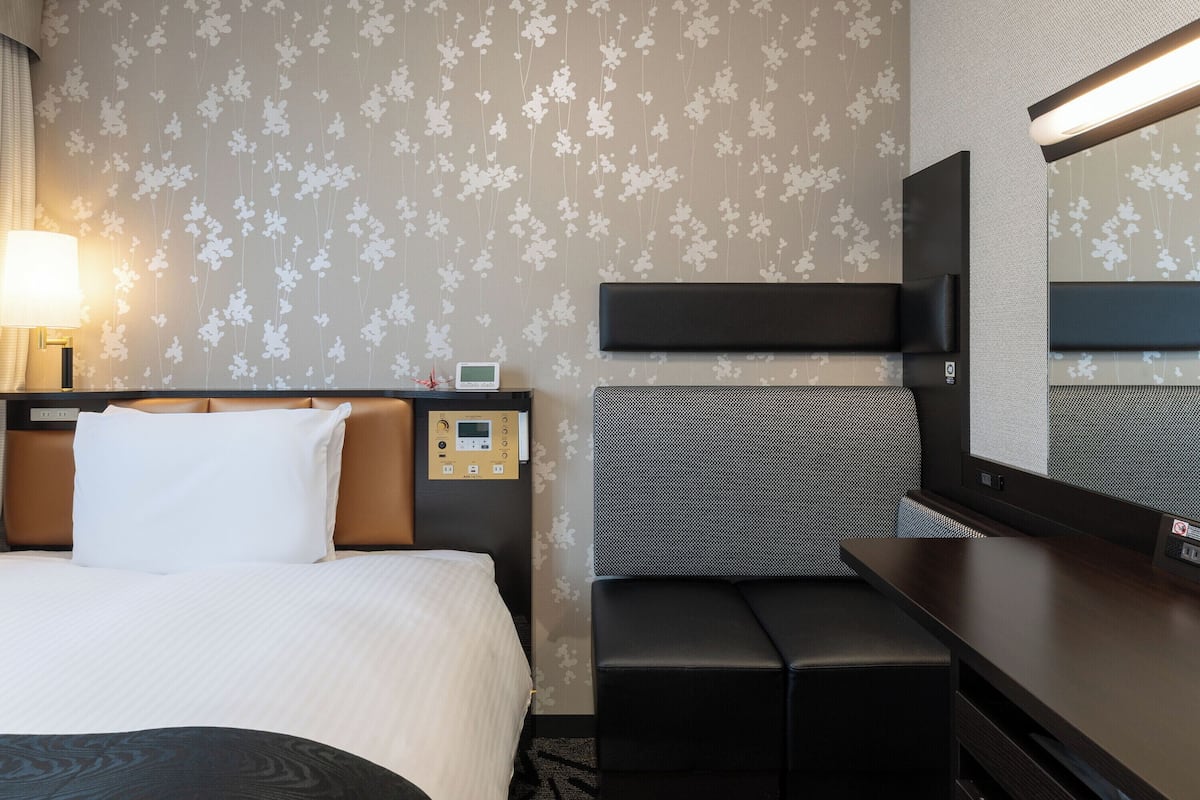 A hotel room with a bed, bedside table, lamp, control panel, bench, and a mirror.