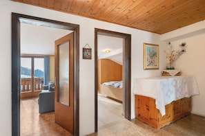 A room with a wooden ceiling, a bed, a small table with a white cloth, a painting on the wall, and a view of a balcony with a chair.