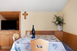 A dining area with a table set for two, a wine bottle, a glass, a cross on the wall, and a flower vase.