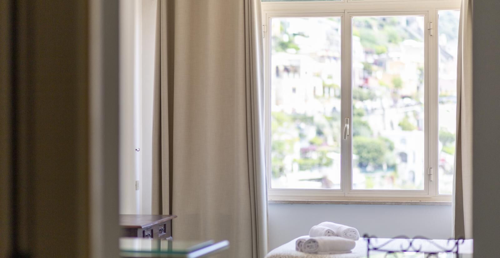 A room with a window, a bed with rolled towels, and a small table with a lamp.