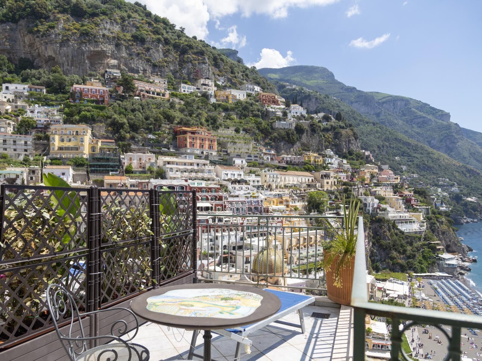 A balcony with a table and chairs overlooking a coastal town with buildings on a hillside.