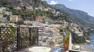 A balcony with a table and chairs overlooking a coastal town with buildings on a hillside.