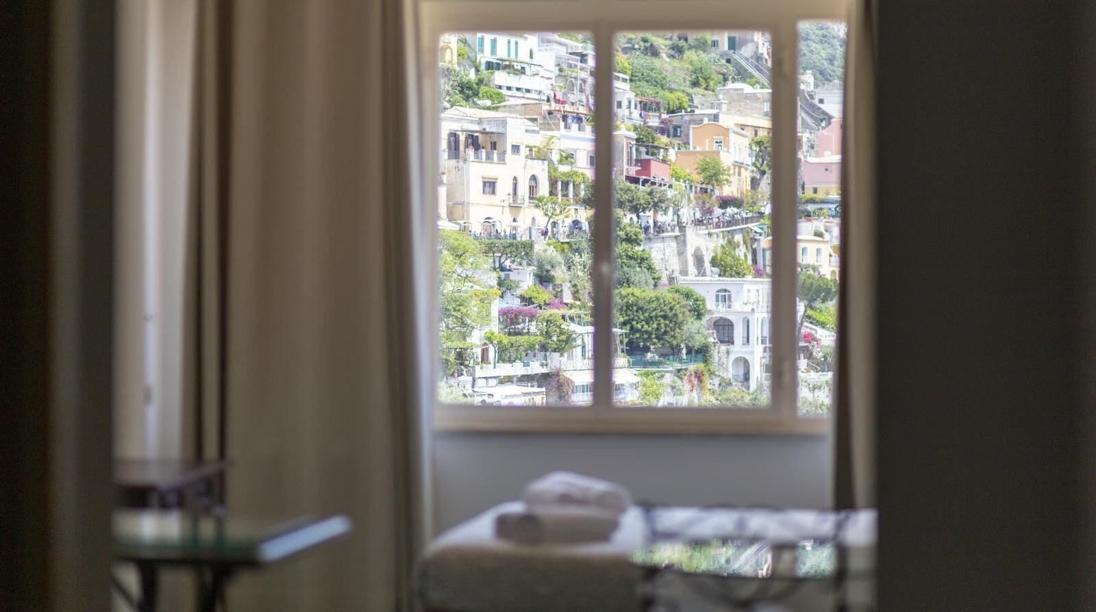 Vista a través de una ventana que muestra un edificio de varios pisos y balcones, una silla y una cama con una colcha blanca.