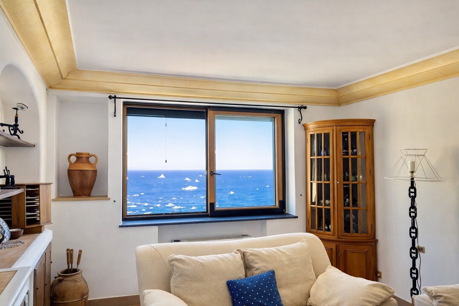 A room with a large window overlooking the sea, a wooden cabinet, and a beige sofa.