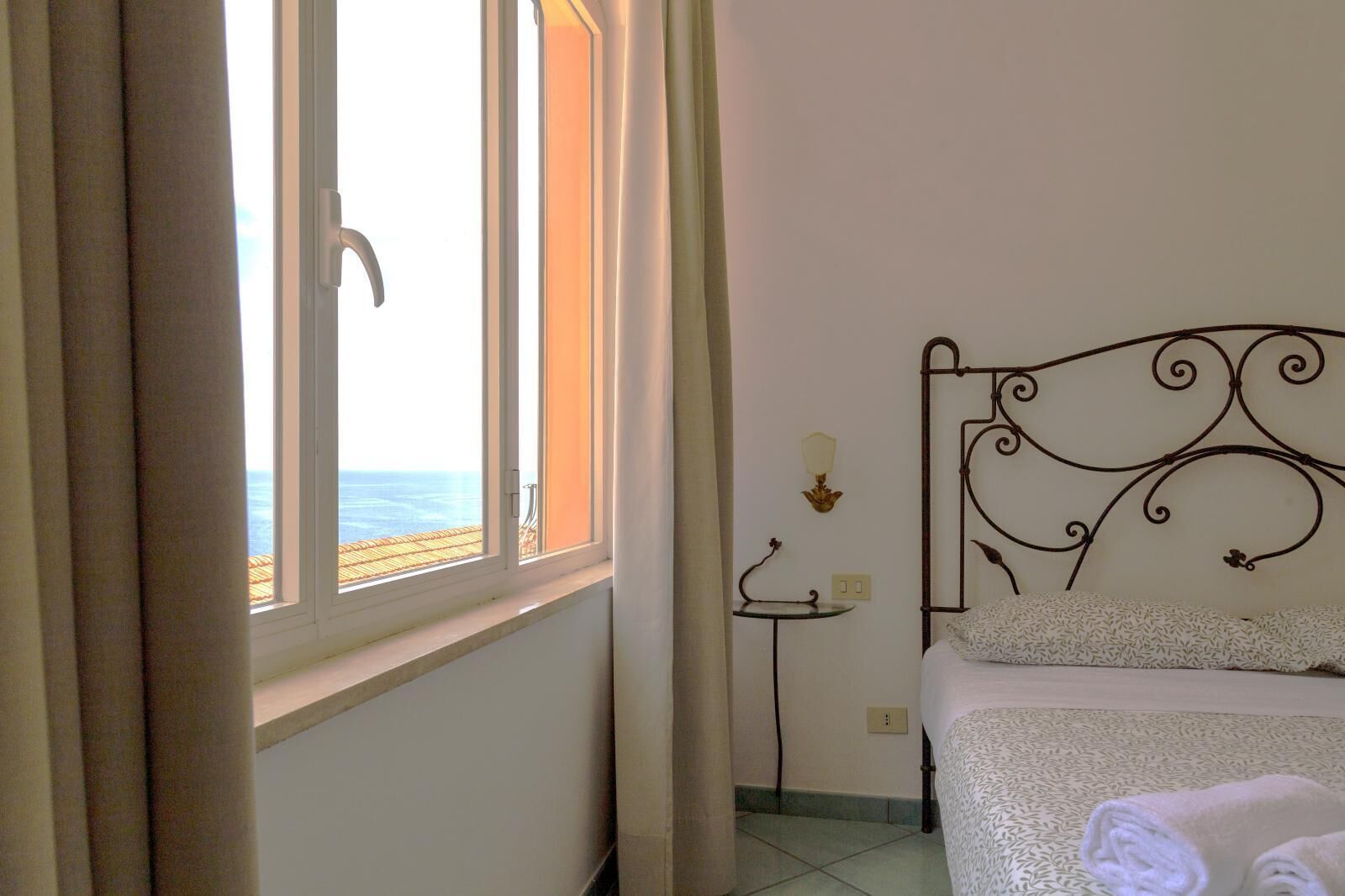 Un dormitorio con cama, mesita de noche, lámpara de pared y ventana con cortinas.