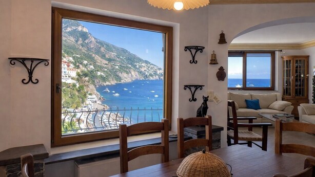 A dining area with a large window overlooking a coastal landscape, wooden furniture, and decorative wall sconces. - Hotiday Positano Sea View (Positano)