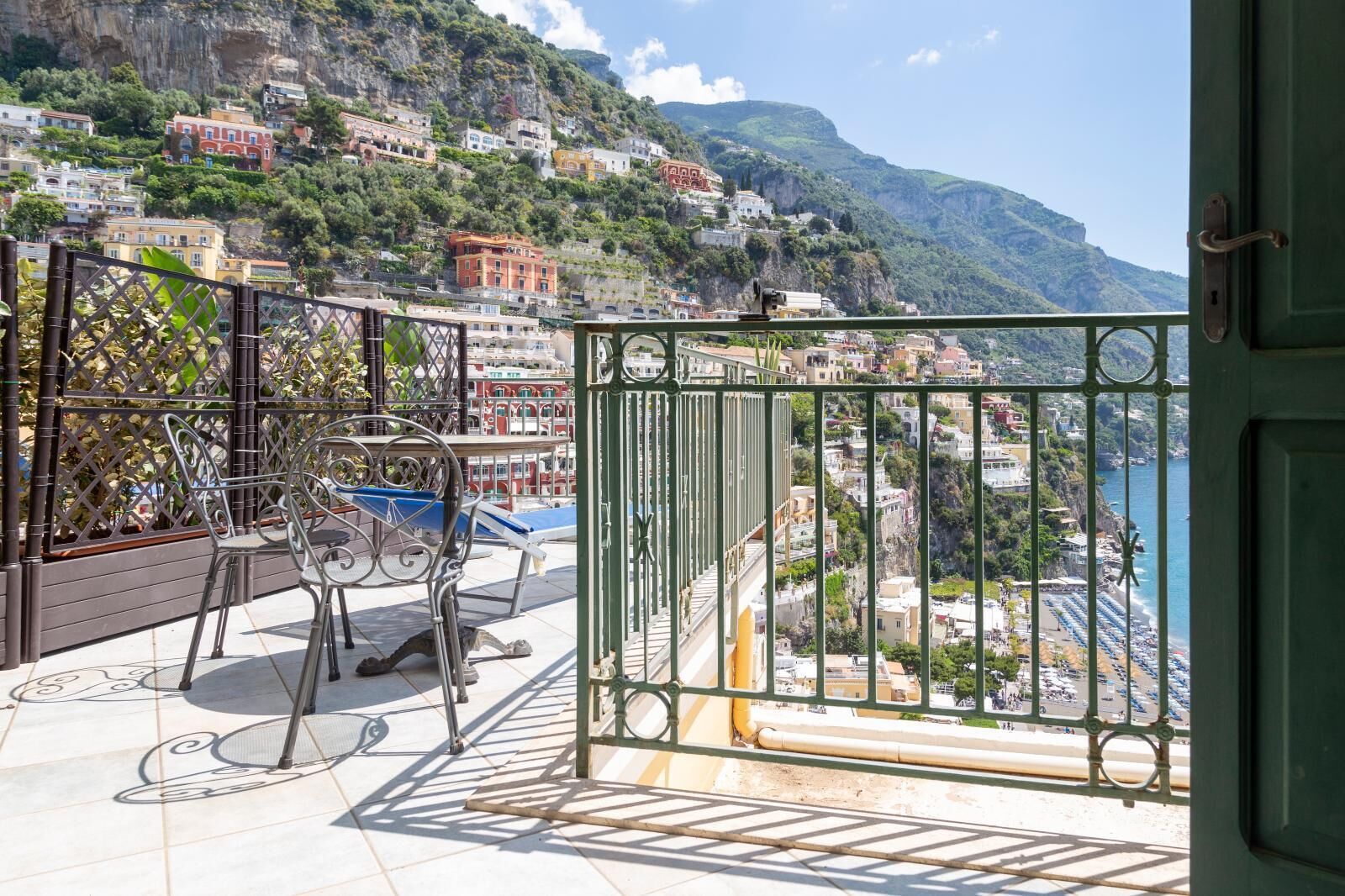 A balcony with a metal railing, a table, and chairs, overlooking a coastal town with buildings on a hillside and a view of the sea.