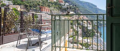 Un balcon doté d’une balustrade en métal, d’une table et de chaises, offrant une vue sur une ville côtière avec des bâtiments construits sur une colline et une vue sur la mer.