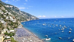 A coastal town with a beach lined with rows of sunbeds and umbrellas, boats in the water, and buildings on a hillside. - Hotiday Positano Sea View (Positano)