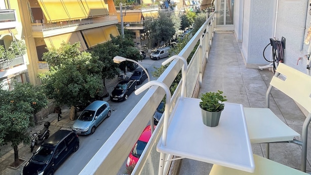 Un balcón con una pequeña mesa y sillas, con vistas a una calle con coches aparcados y edificios.
