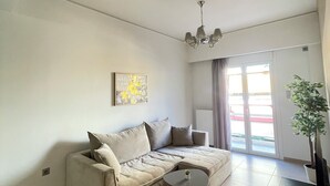 Una sala de estar moderna con un sofá color beige, una mesita redonda pequeña y un sillón a rayas. Se observa una araña, una planta en maceta y un ventanal corredero que da a un espacio exterior.