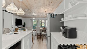 Et moderne køkken med hvide skabe, indbygget ovn, gasblus og kaffemaskine. Der er fletvævet pendellamper, et fladskærms-tv og et spiseområde med bord og stole.