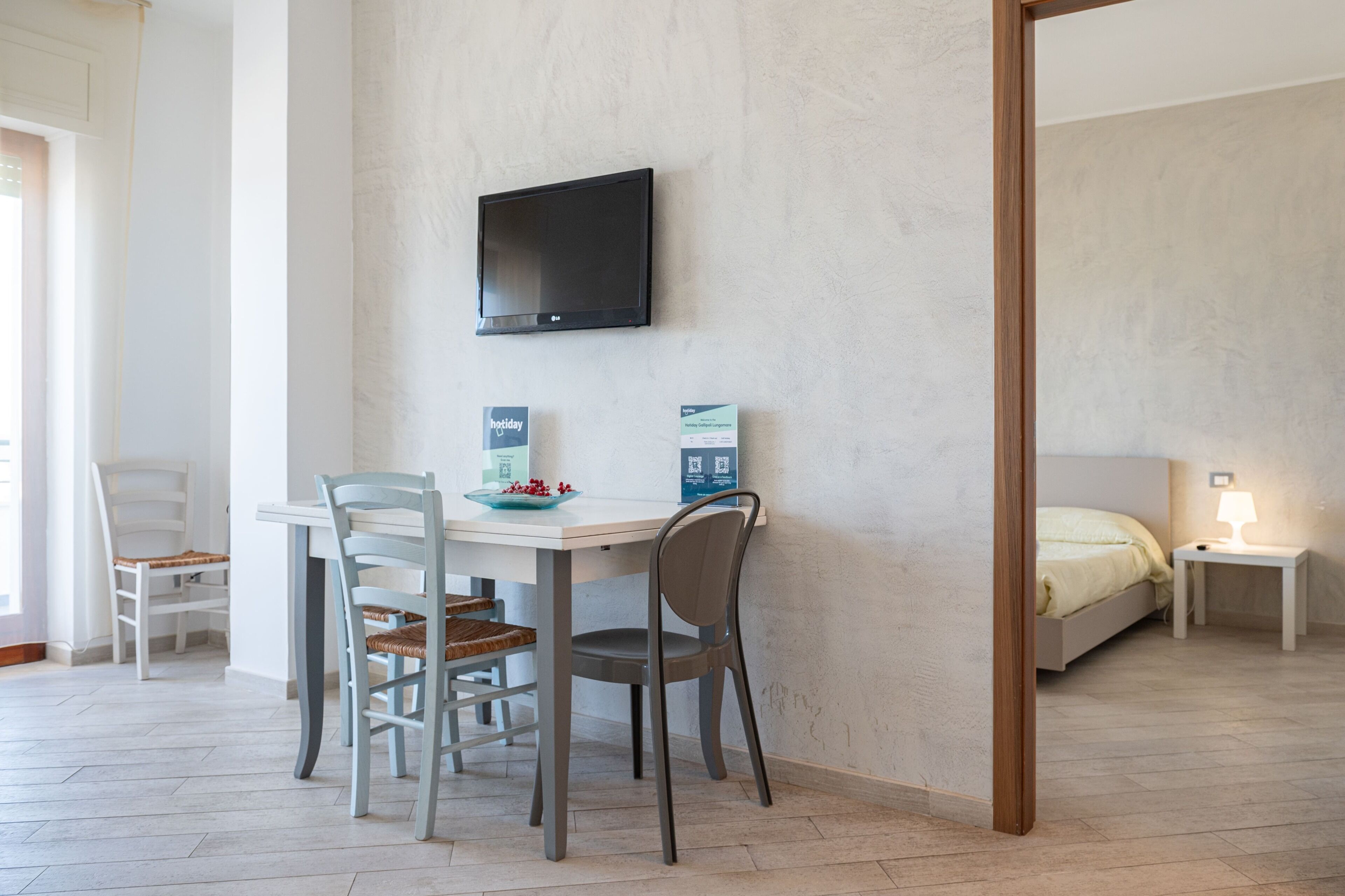 A room with a dining table, chairs, a flat-screen TV mounted on the wall, a bed with a nightstand, and a small table with a lamp.