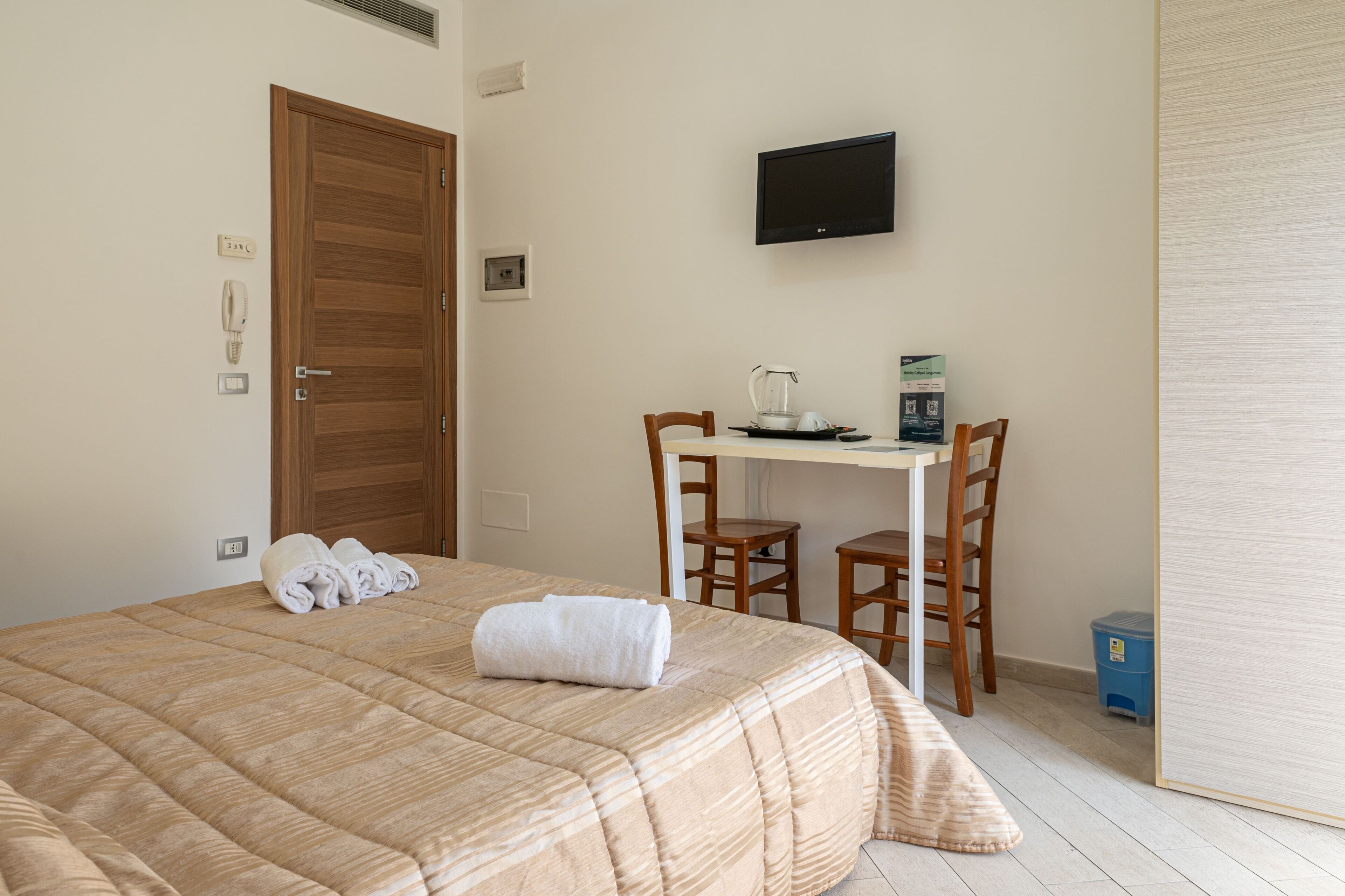 Habitación de hotel con una cama, un escritorio, dos sillas, un televisor y un pequeño cesto de basura.
