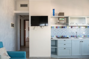 Ett kompakt bostadsutrymme med ett litet kök, en platt-TV som är monterad på väggen, en blå soffa och ett kaklat stänkskydd i köket.