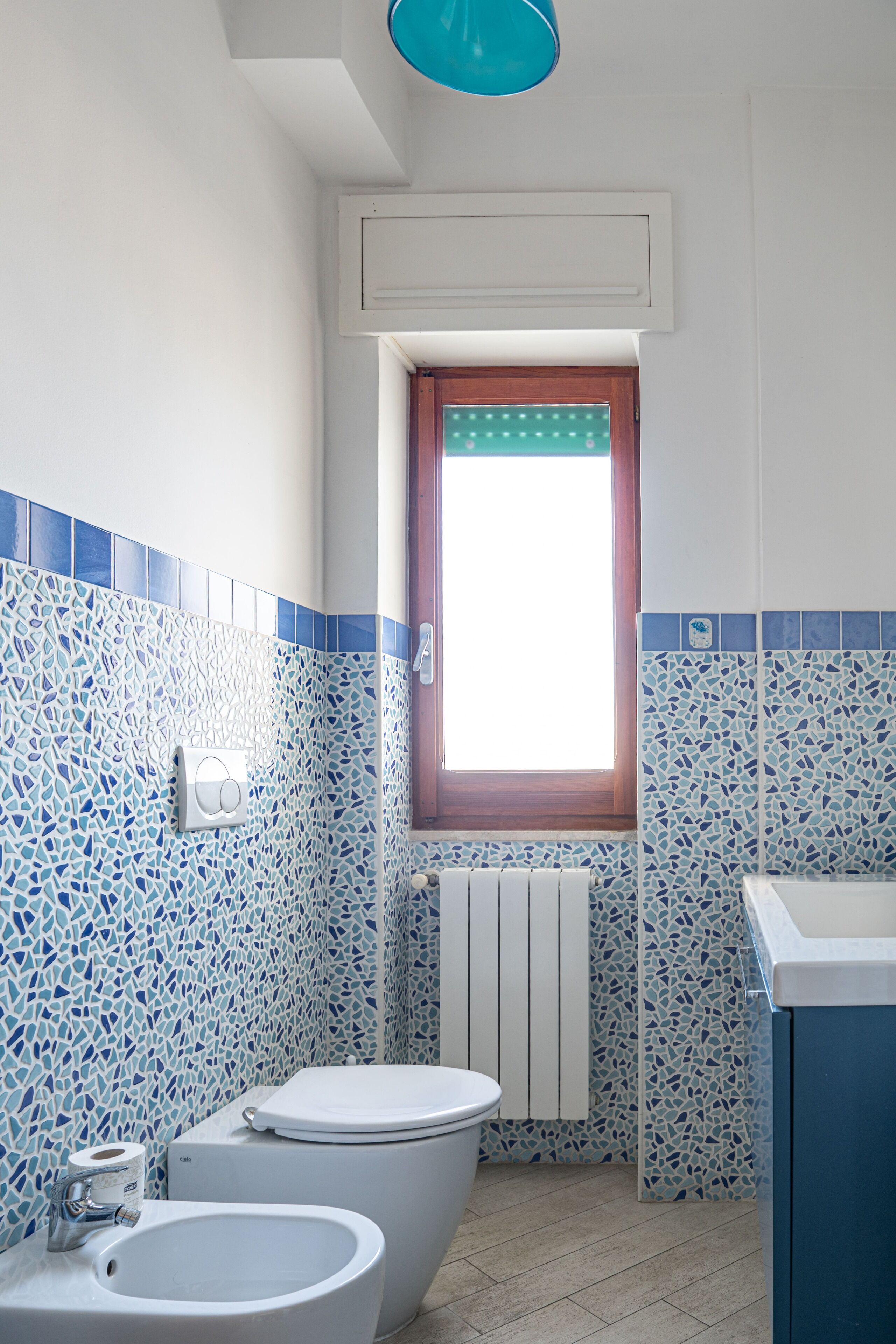 Une salle de bain comprenant des toilettes, un bidet et un lavabo, dont le sol et les murs sont recouverts de mosaïques bleues et blanches, avec une fenêtre située au-dessus des toilettes.