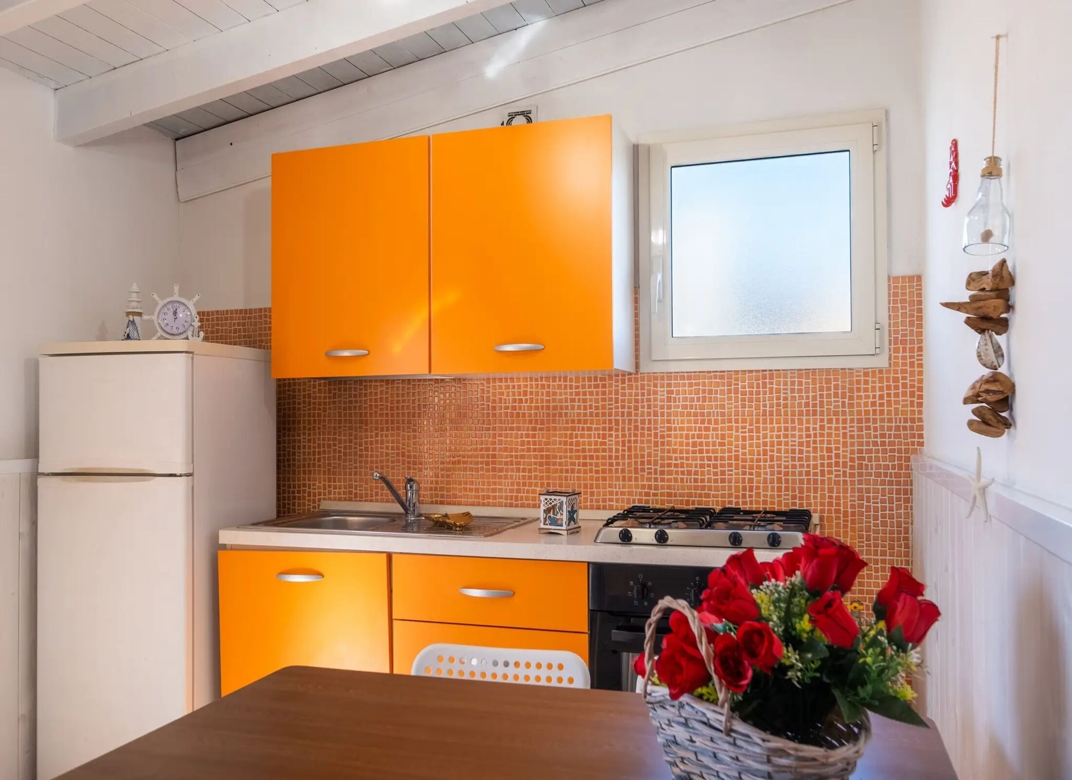 Une cuisine avec des meubles de couleur orange, un réfrigérateur blanc, une cuisinière à gaz et une fenêtre au-dessus de l’évier.