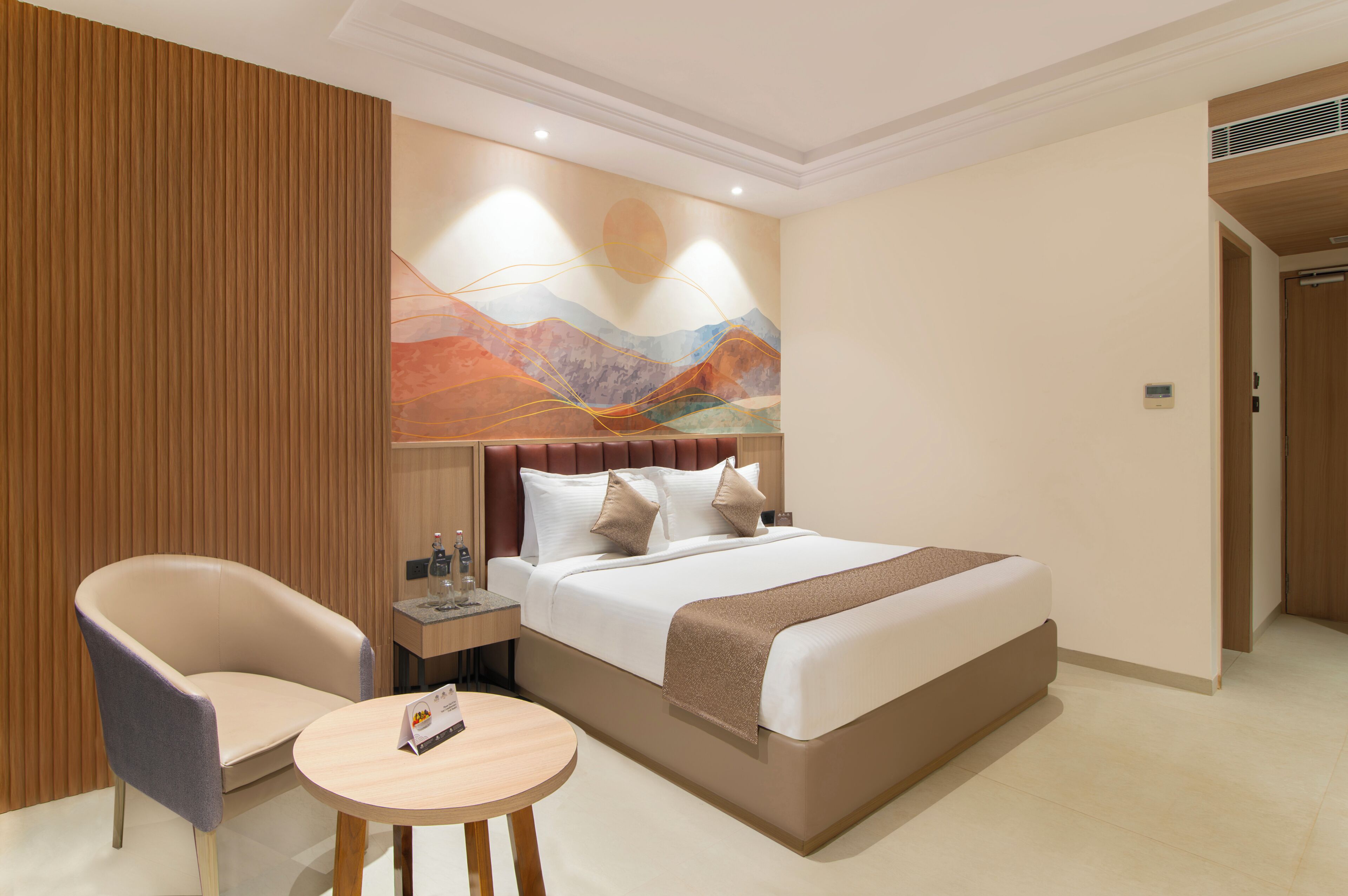 A hotel room with a bed, two chairs, a small table, a nightstand, and a wall art piece.