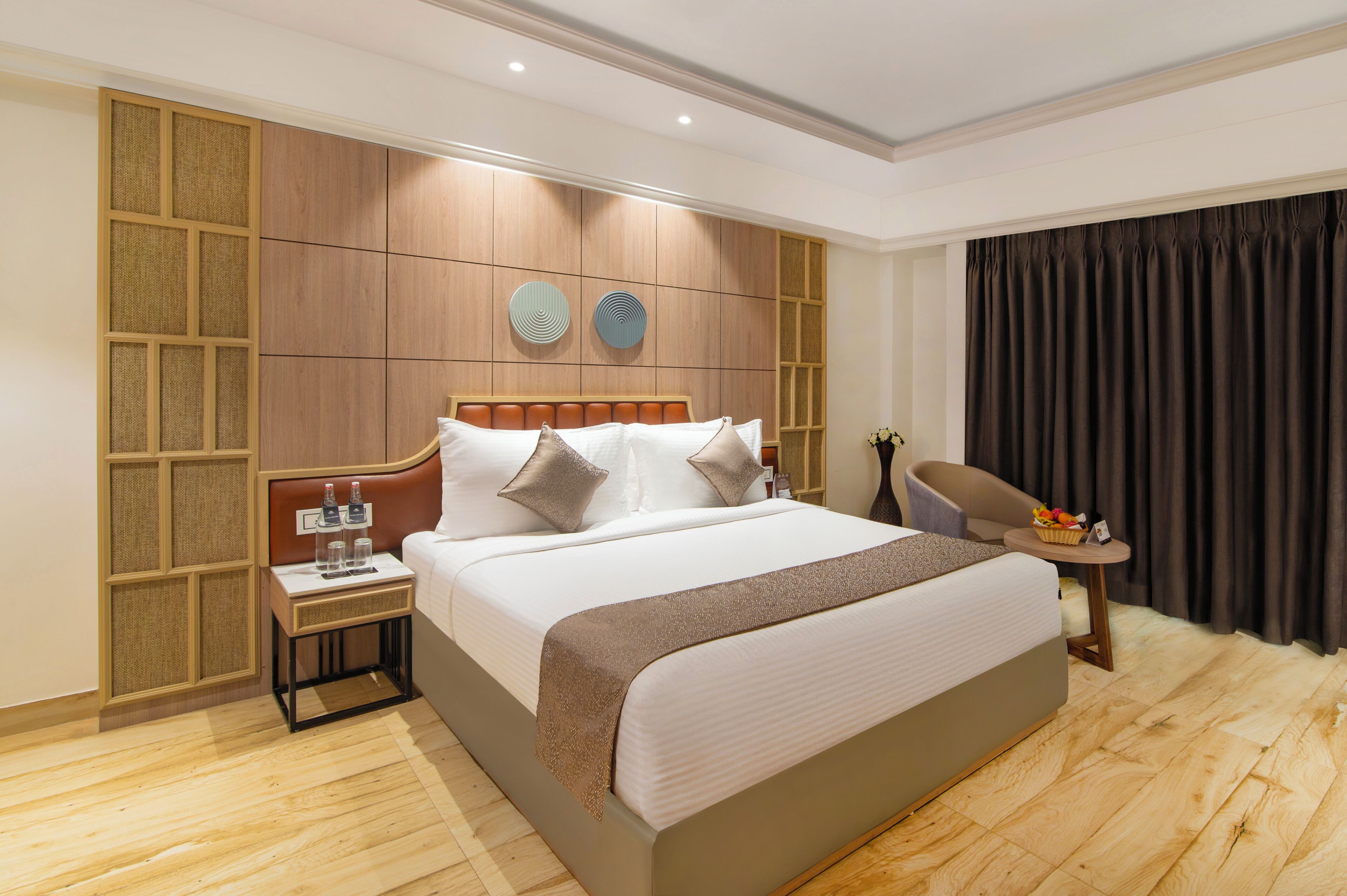 Une chambre d’hôtel comprenant un grand lit, une table de chevet, une chaise, une petite table avec un panier, et un mur orné d’un panneau décoratif et de deux œuvres d’art murales de forme circulaire.