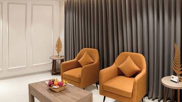 Una habitación con dos sillones naranjas, una mesa de centro de madera con fruta, una mesita auxiliar con un objeto decorativo y un cortinero grande.