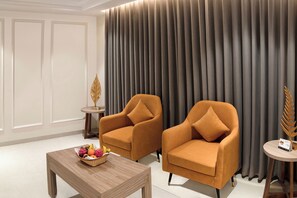 A room with two orange armchairs, a wooden coffee table with fruit, a small side table with a decorative item, and a large curtain. - Enrise By Sayaji Somnath (Veraval)