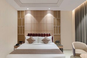 A hotel room with a large bed, a nightstand, a chair, and a wall with a wooden panel design. - Enrise By Sayaji Somnath (Veraval)