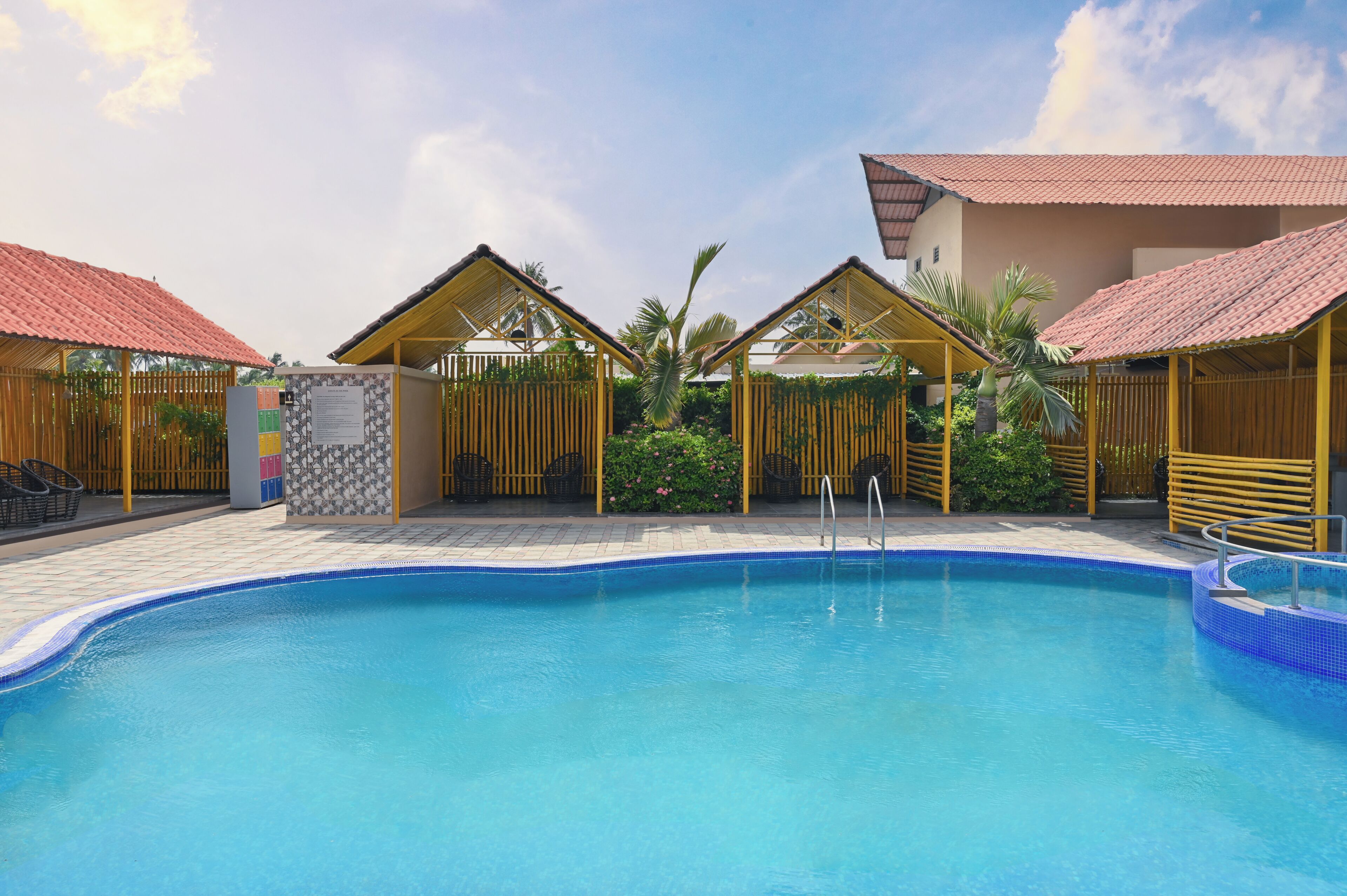 A swimming pool with a tiled edge, surrounded by covered seating areas with red roofs, and a wooden fence with greenery.