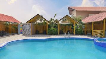 A swimming pool with a tiled edge, surrounded by covered seating areas with red roofs, and a wooden fence with greenery.