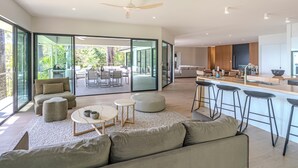 A modern living room with a large sofa, ottomans, and a coffee table. A kitchen area with bar stools and a dining table is visible through a glass door. - NEW Luxury Hamilton Island Estate (Whitsundays)