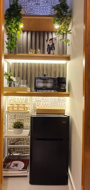 Une cuisine de taille réduite, avec un réfrigérateur noir, un four à micro-ondes et une étagère en bois au-dessus.