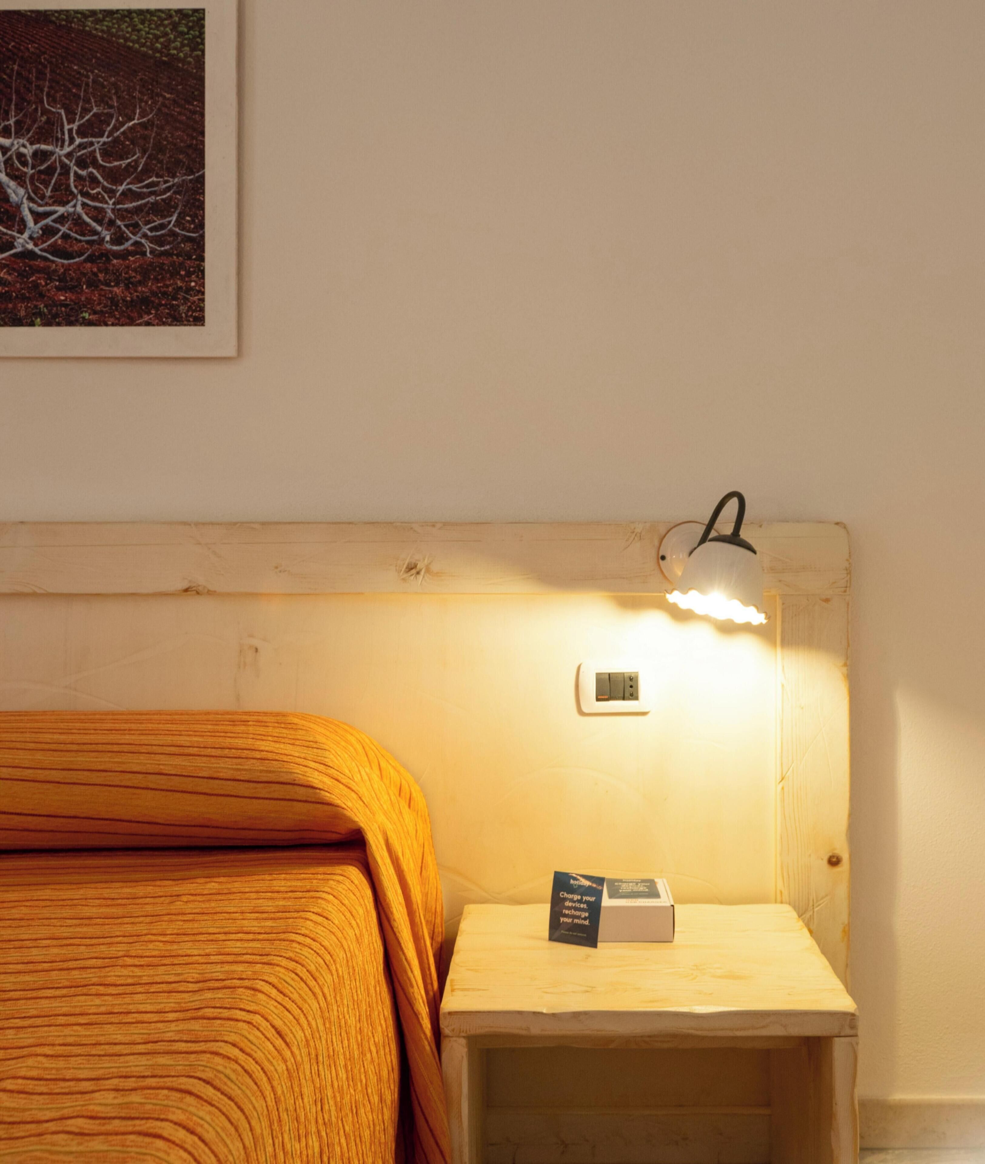 Una cama bien hecha con cabecera de madera, una mesita de noche con una lámpara y una tarjeta pequeña sobre la mesa.