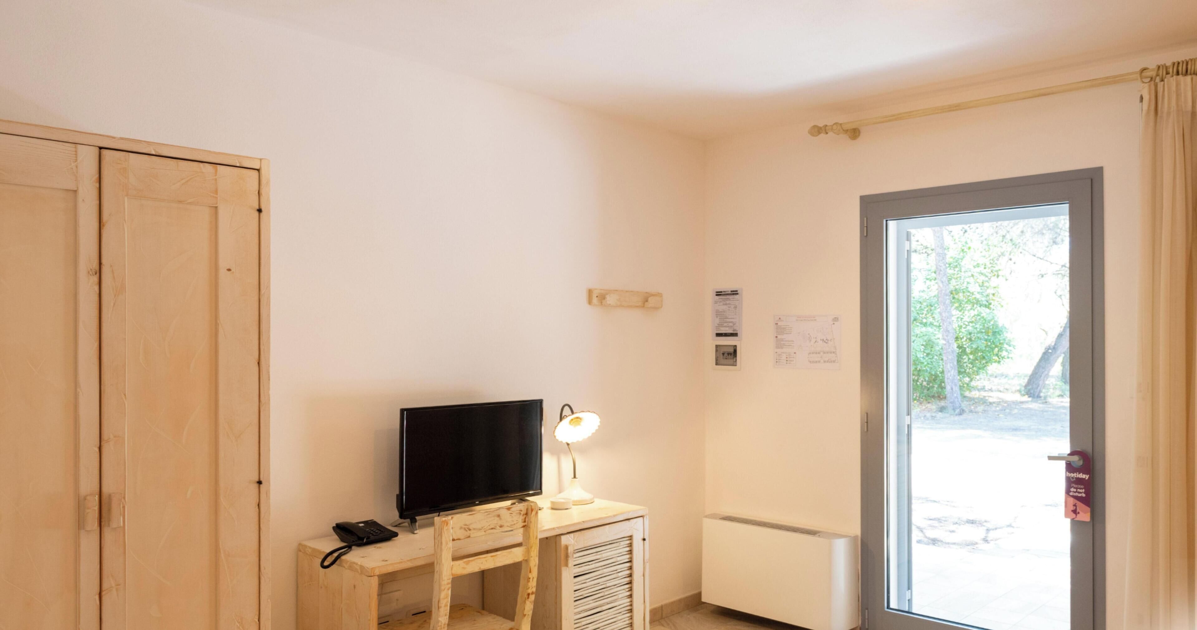 Ένα δωμάτιο με μια τηλεόραση πάνω σε ένα ξύλινο γραφείο, ένα φωτιστικό, ένα καλοριφέρ και μια πόρτα από την οποία φαίνεται η θέα προς τα έξω.