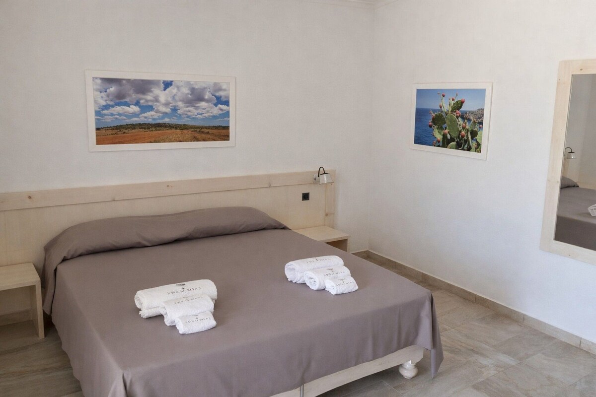 Een slaapkamer met een bed, twee handdoeken, een nachtkastje, een afbeelding van een woestijnlandschap en een afbeelding van een cactus aan zee.