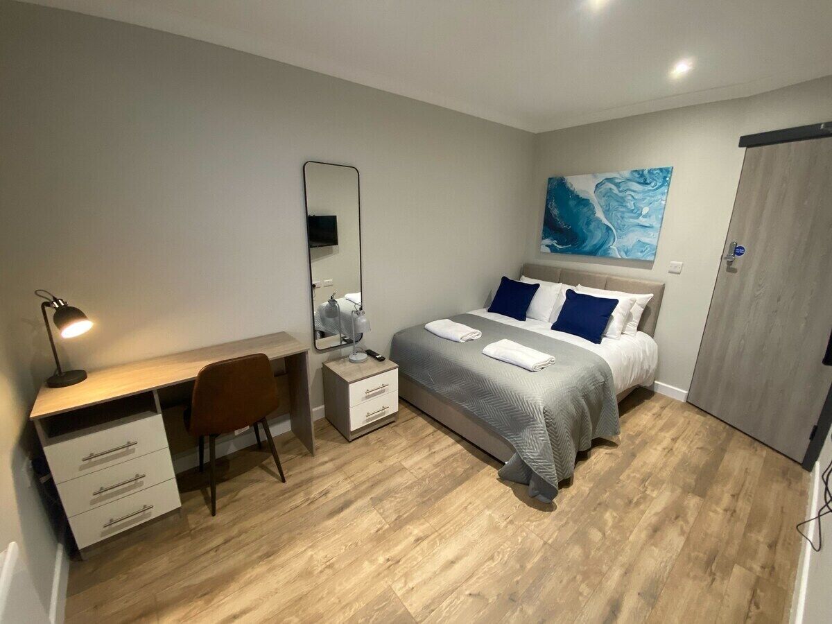 A bedroom with a bed, a desk, a chair, a dresser, a mirror, and a painting on the wall.