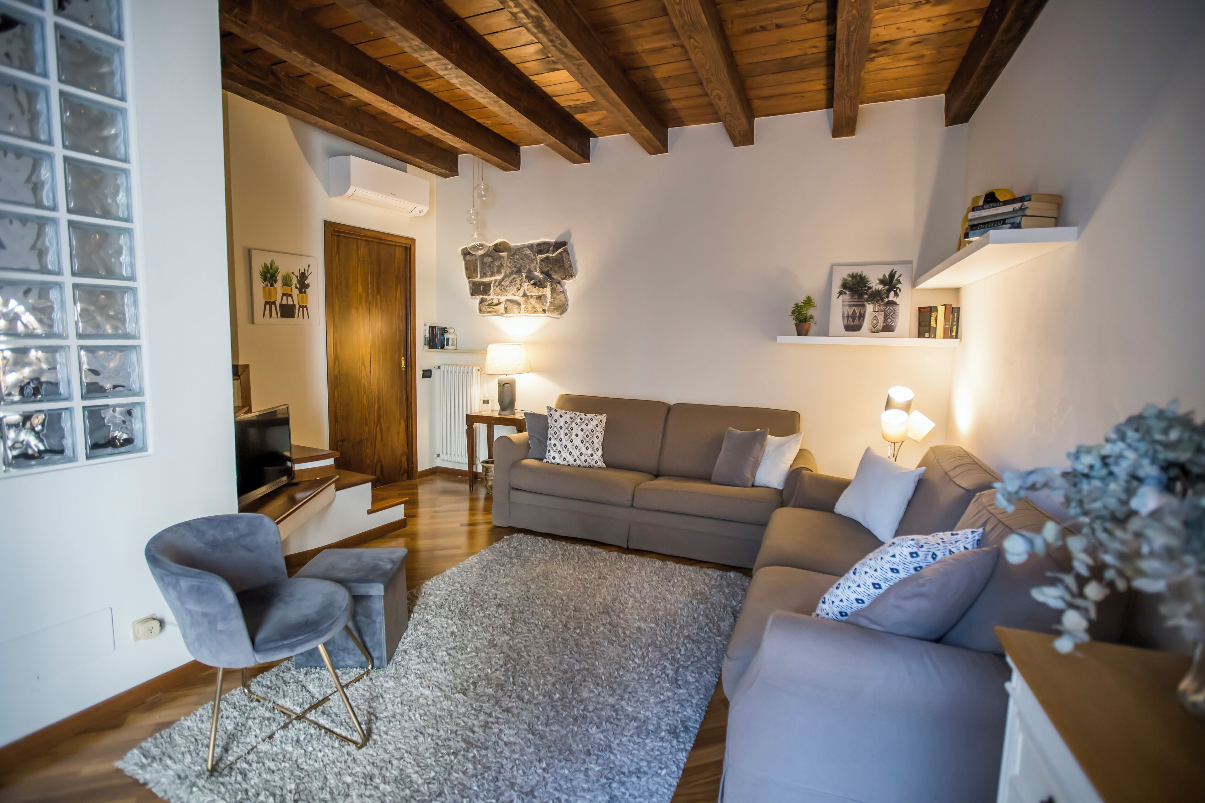 A cozy living room with a sofa, armchairs, and a rug. There are wall-mounted shelves with decorative items and a wooden ceiling.