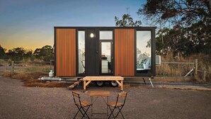 Une petite cabane moderne dotée d’une porte coulissante, d’une table et de deux chaises à l’extérieur.