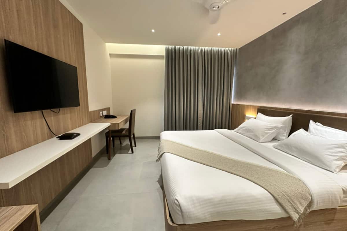 A modern hotel room with a flat-screen TV, a desk, a chair, a bed with white linens, and a large window with curtains.