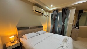 A hotel room with a bed, bedside lamp, air conditioning unit, and a view of the outside through curtains. - Lexis Residence - Newly Renovated Rooms (Tambon Patong)