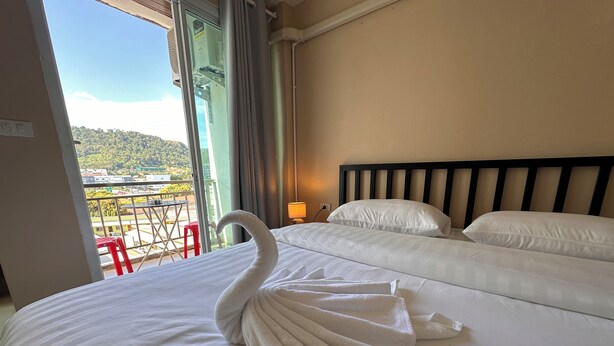 A hotel room with a bed, a swan-shaped towel, a balcony with a red chair, and a view of a green landscape. - Lexis Residence - Newly Renovated Rooms (Tambon Patong)