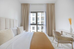 A hotel room with a large bed, a desk, a chair, and a view of a building through the window. - Bloomfields Cozy 1br Apt Town (Dubai)