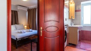 A room with a bed, a wooden door, a washing machine, and a bathroom with a mirror and sink. - Scala Nostra Apartment (Corfu)