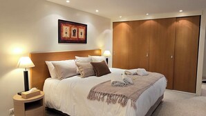 Ein modernes Schlafzimmer mit einem großen Bett, einem Nachttisch mit Lampe, einem Holzschrank und einem gerahmten Bild an der Wand.