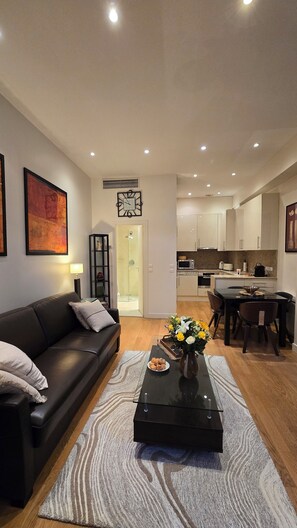 A modern living room with a black sofa, a coffee table with a vase of flowers, and a kitchen in the background. - Chic Parisian Retreat Near Louvre, Cafés & Couture (Paris)