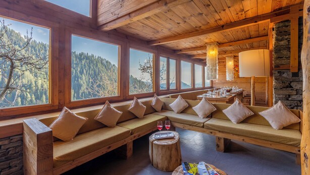 A wooden interior with large windows, a stone wall, and a wooden bench with cushions. - ELIVAAS Silvana 3 BHK Cottage  (Kumharsain)