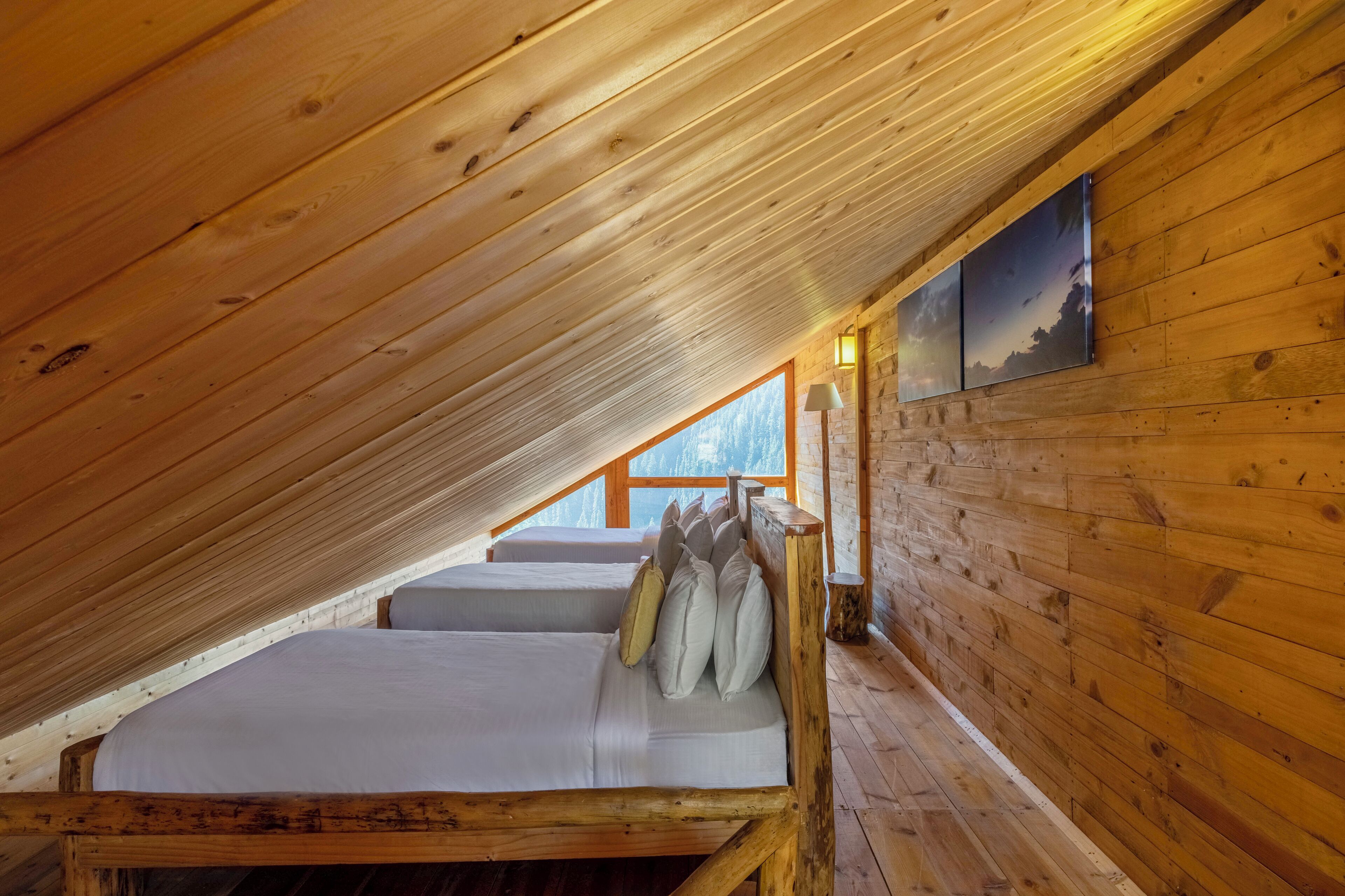 A cozy attic bedroom with wooden walls and a sloped ceiling. There is a bed with pillows and a nightstand with a lamp. A painting hangs on the wall, and a window provides a view of the outdoors.