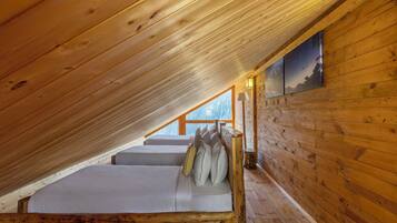 A cozy attic bedroom with wooden walls and a sloped ceiling. There is a bed with pillows and a nightstand with a lamp. A painting hangs on the wall, and a window provides a view of the outdoors.