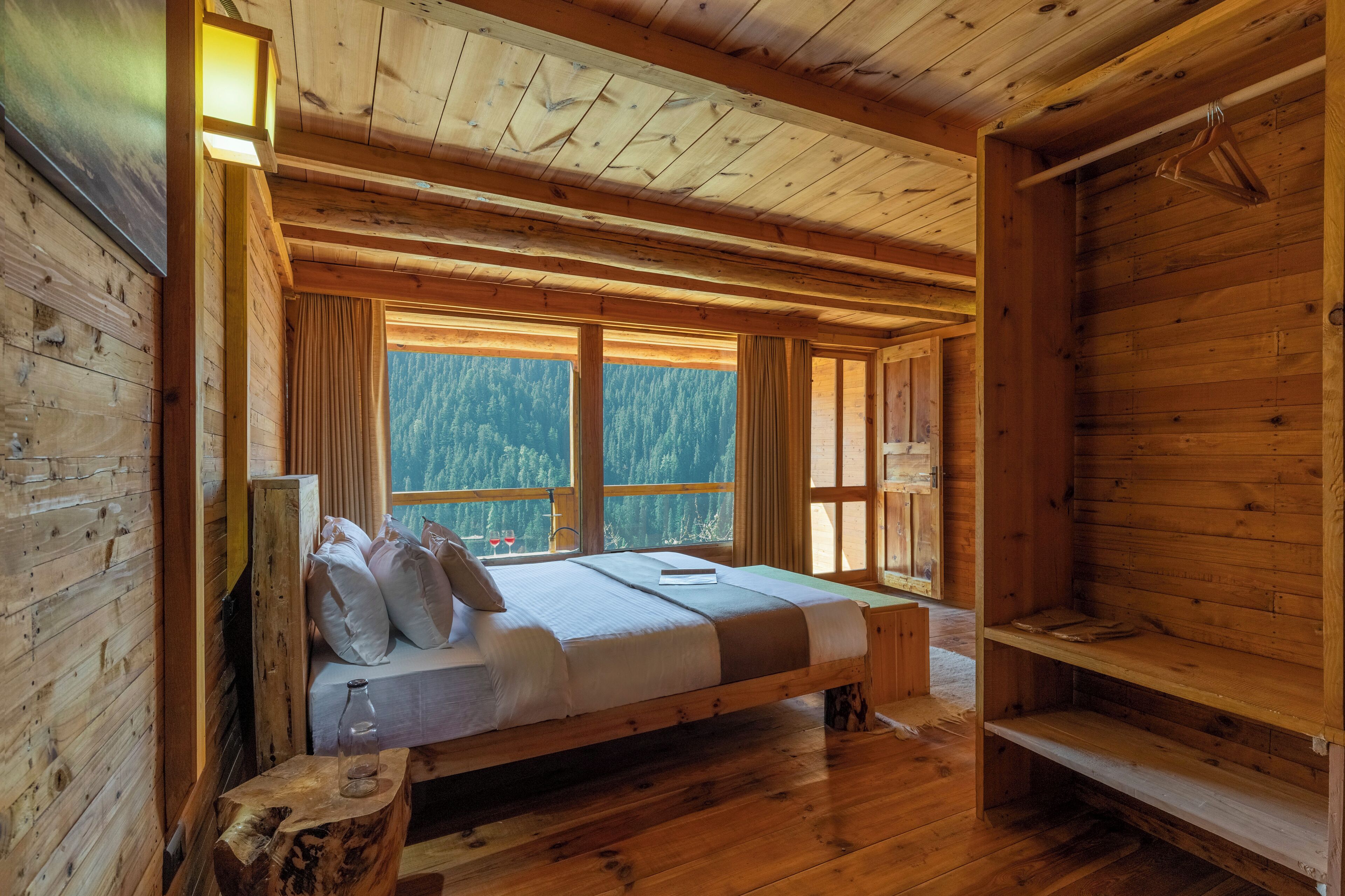 Ein Schlafzimmer in einer Holzhütte mit einem Bett, einer Bank und einem Fenster mit Blick auf einen Wald.