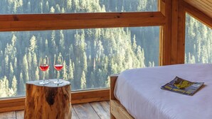 A bedroom with a large window, a wooden bed, and a small table with two glasses of red wine. - ELIVAAS Silvana 3 BHK Cottage  (Kumharsain)